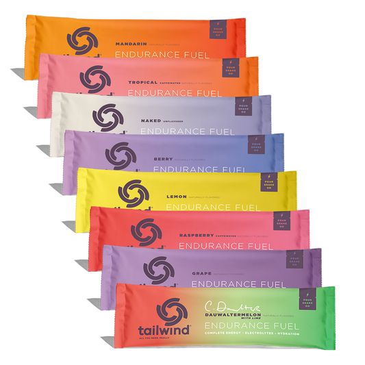 Eight colorful Tailwind Endurance Fuel packets are stacked, displaying a range of flavors like Mandarin, Tropical, and Dauwaltermelon.