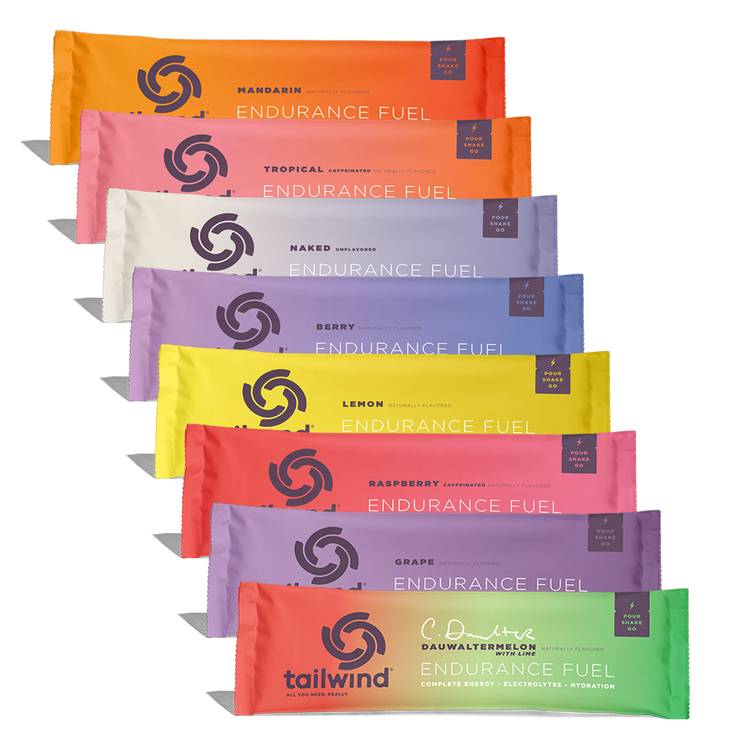 Eight colorful Tailwind Endurance Fuel packets are stacked, displaying a range of flavors like Mandarin, Tropical, and Dauwaltermelon.