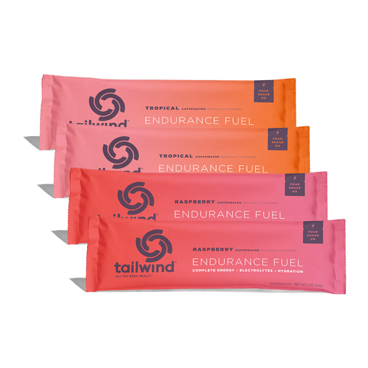 Studio product photography by Tailwind Nutrition of four staggered Endurance Fuel stick packs in a central studio arrangement featuring vibrant orange and magenta color gradients, dark blue circular swirl logos, and labels for tropical and raspberry caffeinated mixes