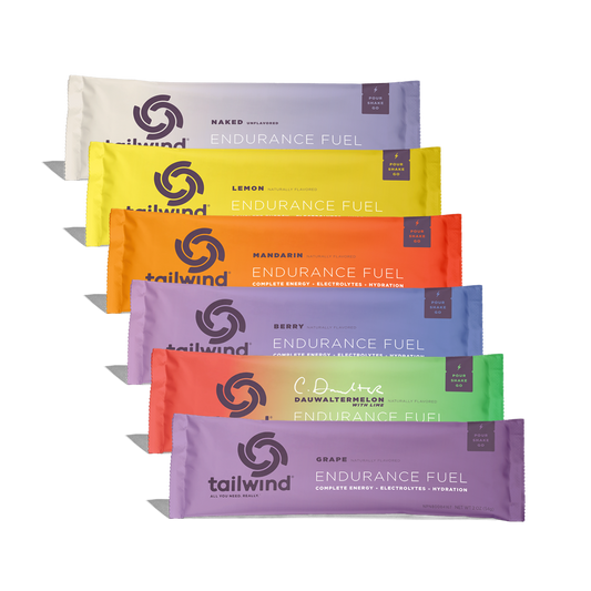 Studio product photography of Tailwind Endurance Fuel packets stacked diagonally against a solid black background, showcasing vibrant colors like lemon yellow and mandarin orange alongside the brand signature circular whirlwind logo and Dauwaltermelon flavor packet