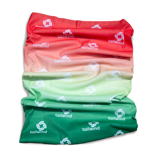 Product photograph by Tailwind of a neck gaiter on a black background featuring a vibrant red-to-green ombre gradient and repeated white Tailwind logos on lightweight pleated fabric