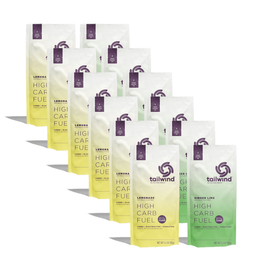 Commercial product photography by Tailwind Nutrition of High Carb Fuel drink mix packets in yellow and green flavor gradients featuring a purple swirl logo and 90g carbs badge arranged in two rows against a black background