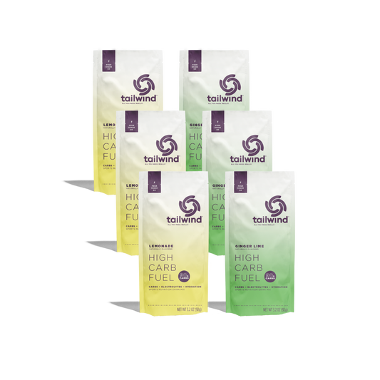 Product photograph of Tailwind Nutrition showing six High Carb Fuel packets on a black background featuring three bright yellow Lemonade and three pale green Ginger Lime pouches with purple spiral logos and circular 90g carb badges