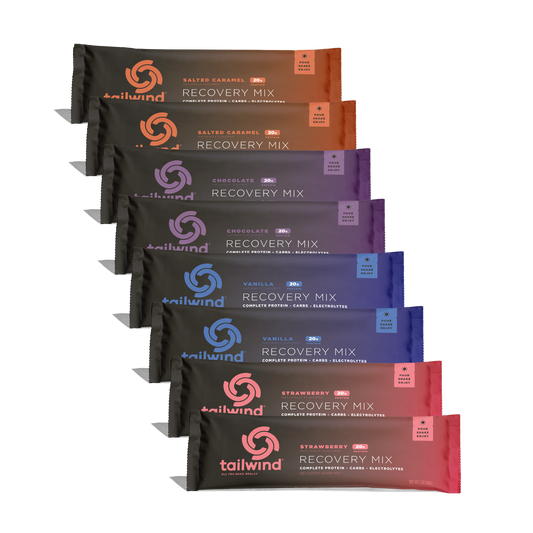 Product photography by Tailwind Nutrition featuring a diagonal stack of eight Recovery Mix packets on a black background showcasing the brand’s interlocking circle logo alongside vibrant orange, purple, blue, and pink flavor accents and 20g protein labels